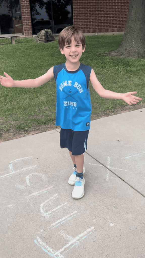 young boy dressed in blue athletic clothes smiles at the camera with his arms wide showcasing the obstacle course drawn with chalk on the sidewalk.