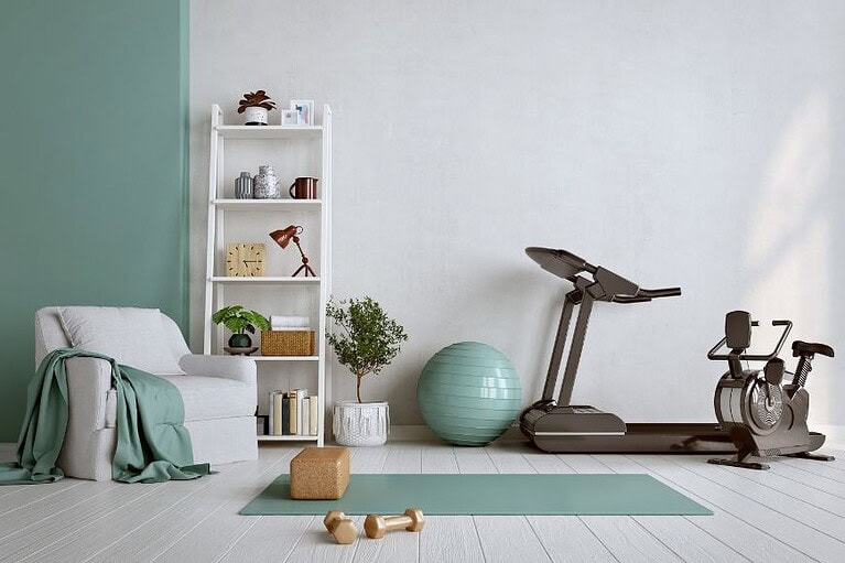an aqua blue yoga mat, walking pad, free weights and a blue ab ball sits in an home gym. white wall with bookshelf showcasing home accessories