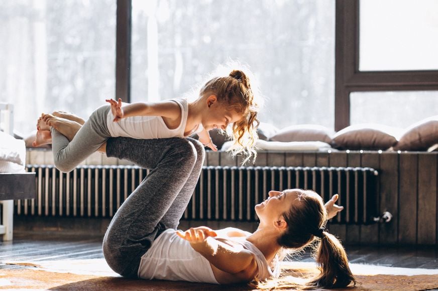 mom on the floor on her living room playing "airplane" a yoga post that holds her daughter on her shines high in the air.