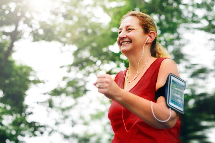 woman wearing a red athletic top running outdoors and smiling. she has her phone strapped to her arm