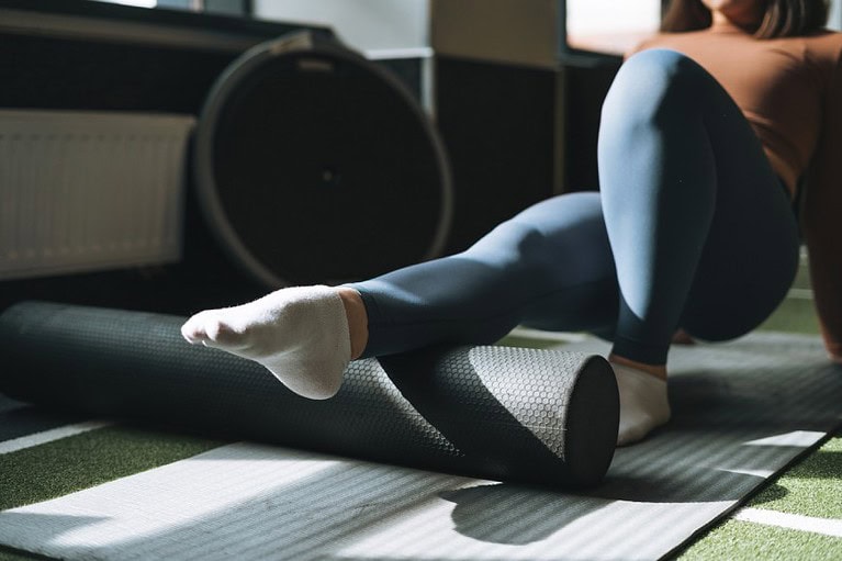 a woman wearing blue athletic pants and white socks shown foam rolling her calf muscle with one leg pointed and one leg bent. the foam roller is dark grey as is the yoga mat she is shown hovering over