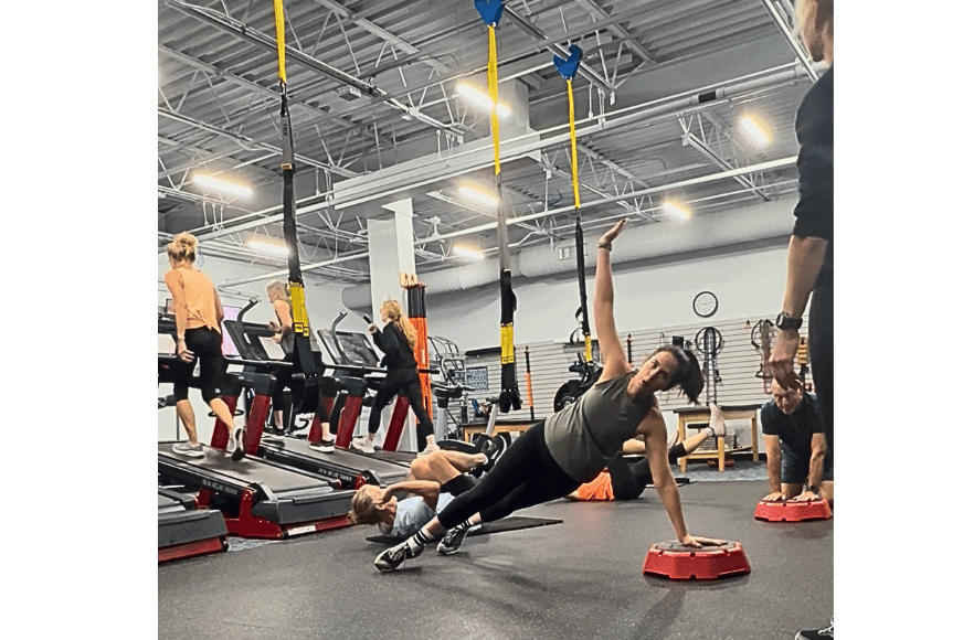 natalie kalmar, fitnessfoodiestl, shown at nutriformance at hike & hybrid group fitness class. she is holding herself up with one arm while other class participants are shown on the treadmill and doing crunches