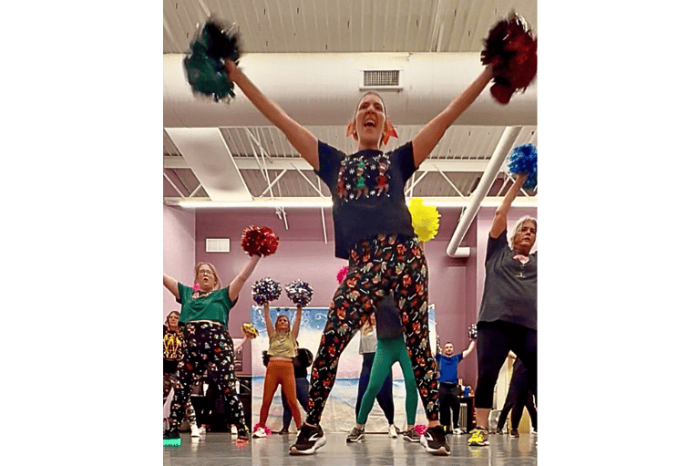 The front of a Pom Squad dance fitness class, where instructor demonstrates a high vee dance move at pom squad fitness dance move and the class participants all with different colored poms follow along.