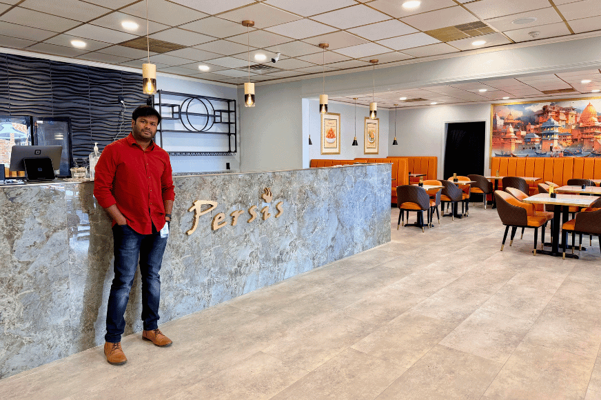 one of the owners of Persis Indian Grill in Ballwin smiling at the front of the restaurant where Persis is written on the stone clad entryway