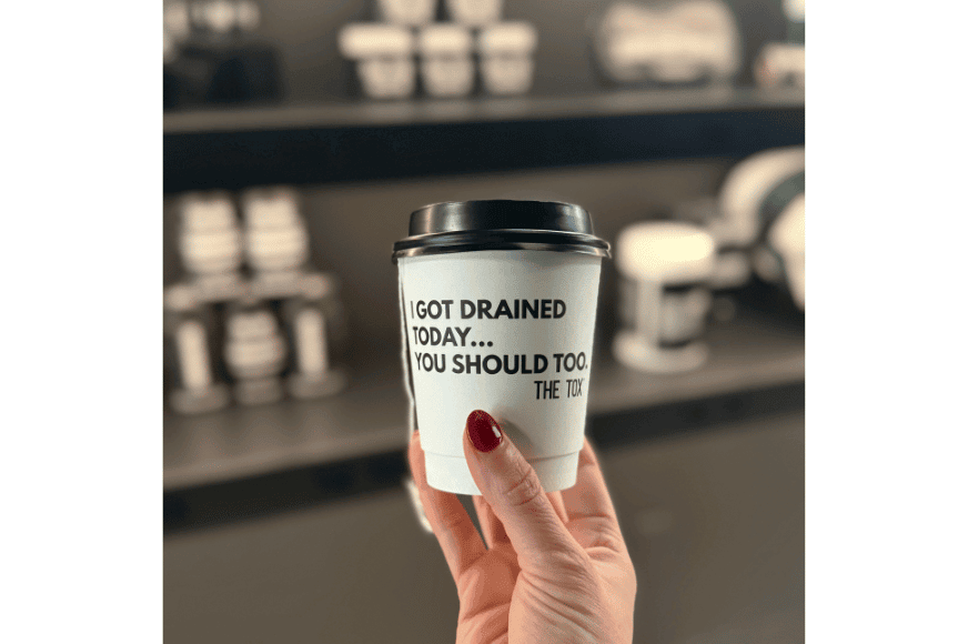 a womans hand holding a white paper cup with black lid and black writing that says "i got drained today, you should too"