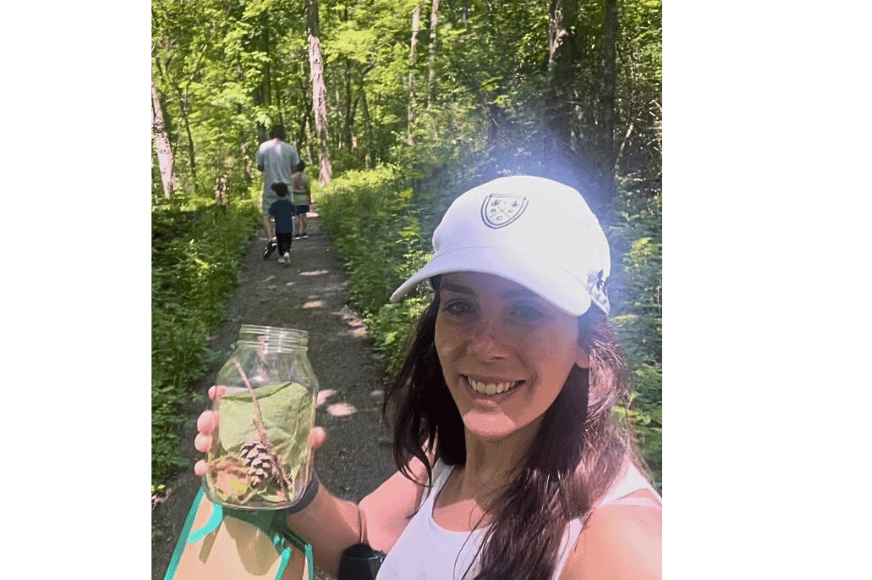 natalie kalmar in a sunny forest on a trail with her kids holidng a glass jar with outdoor trinkets collected like leaves, twigs, rocks and more