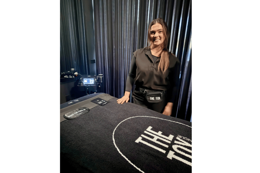 riley an estetitian from the tox is shown standing smiling in front of a treatment table. the room, the table and her outfit is all black.