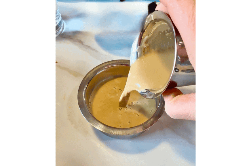 an indian coffee delight, a creamy, light tan colored coffee is being poured into a silver drinking bowl