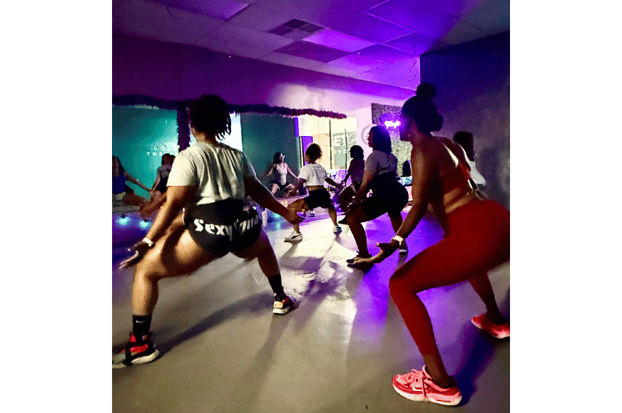 a dark group fitness room with purple lighting and women shown twerking for fitness in short shorts