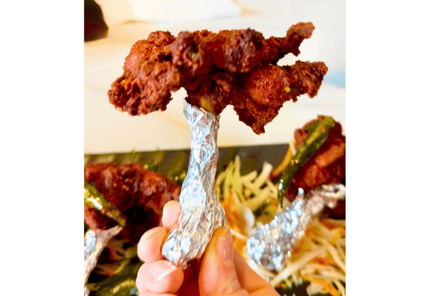 an indian chicken wing known as a chicken lolipop is being held up by its foil base; red & charred splayed for easy biting