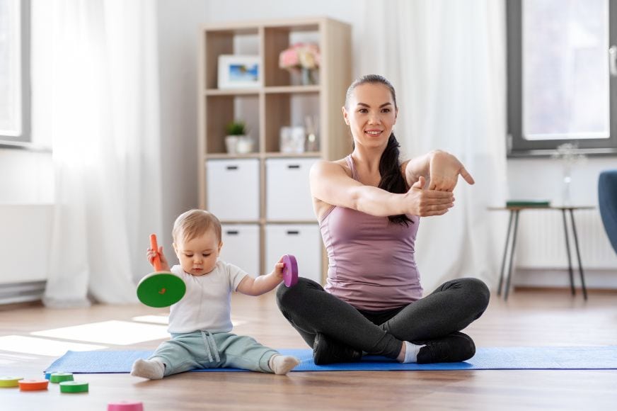 women sitting in her living room on a yoga mat with her toddler next to her strentching out her wrist and bicep while the toddle plays with a toy