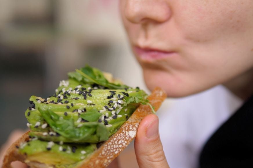 zoomed in photo of a women's mouth that just took a bite of avocado toast - a piece of toast topped with avocado slices and sesame seeds