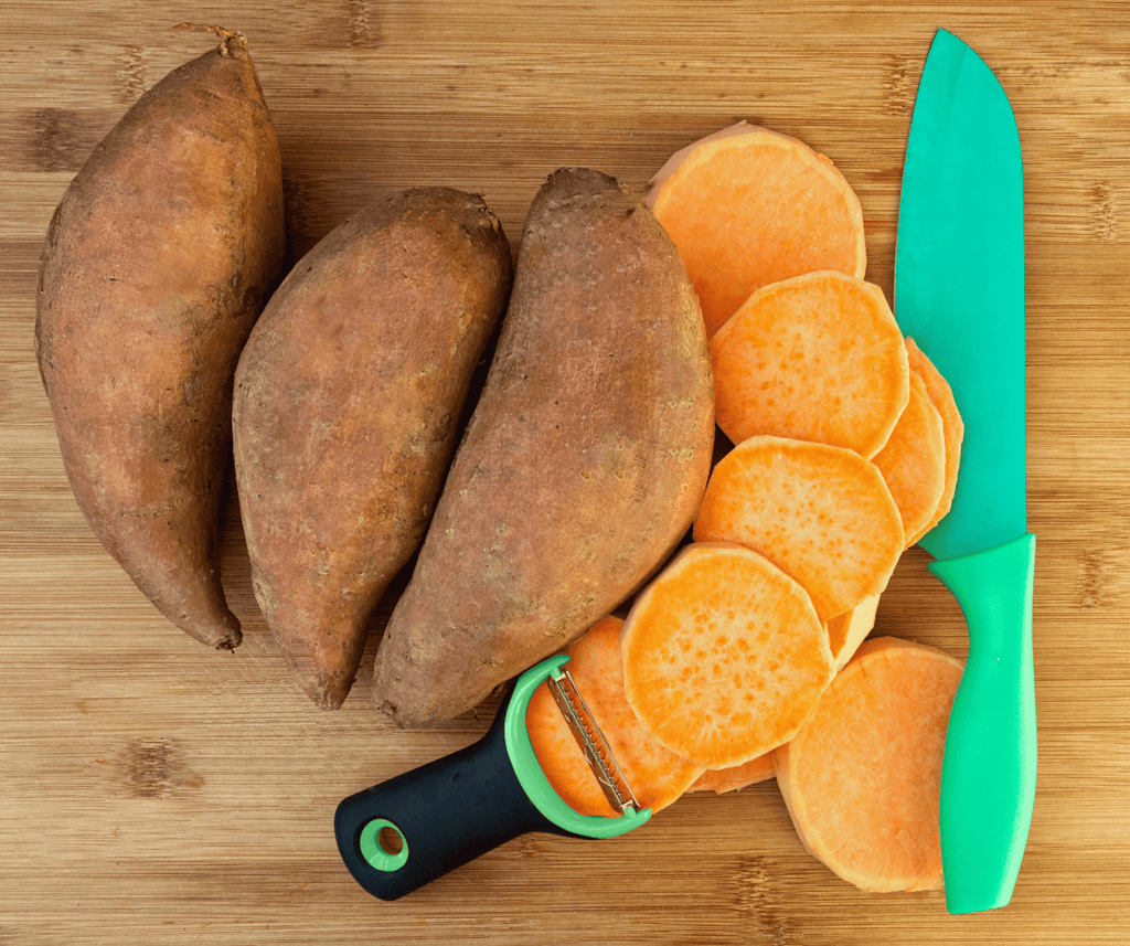 3 unpeel sweet potatoes next to another peeled sweet potato cut into rounds that are fanned on a wooden cutting board next to a green knife and peeler.  