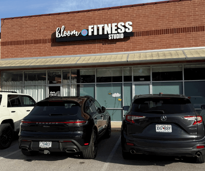 exterior of bloom fitness studio shows class windows with a few cars parked out front of this strip mall dance studio. the bloom logo in the brick exterior wall.