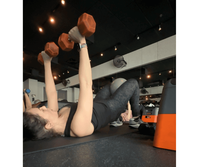 natalie kalmar is shown doing a chest press with orange weights from the floor while squeezing a ball between her knees