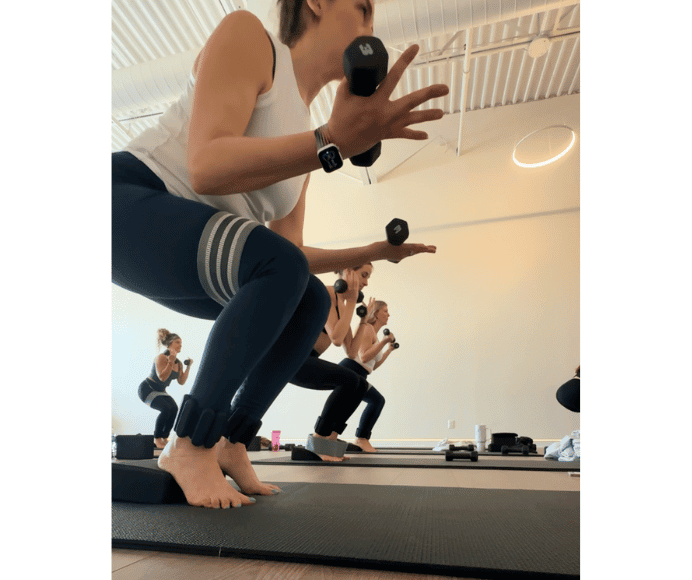 natalie kalmar in a deep squat with a resistance band around her thighs while holding 3lb weights in a class setting