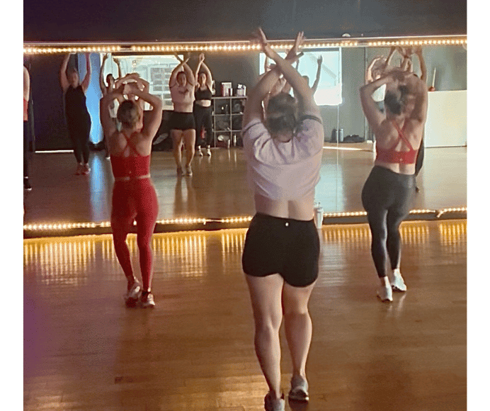3 women wearing bright colored athletic clothes shown dancing with their hands in the air and one hip out while their reflections shines in the mirror