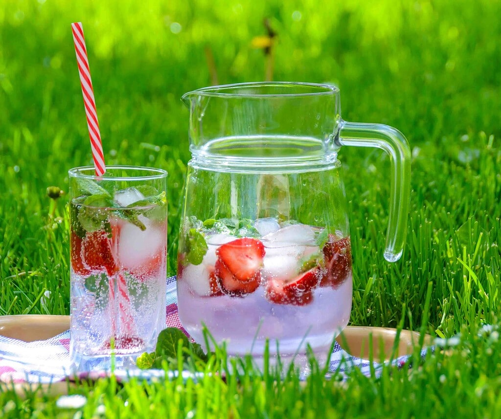 a pitcher of bubbly water with fresh strawberries and a glass filled and a straw set in bright green grass