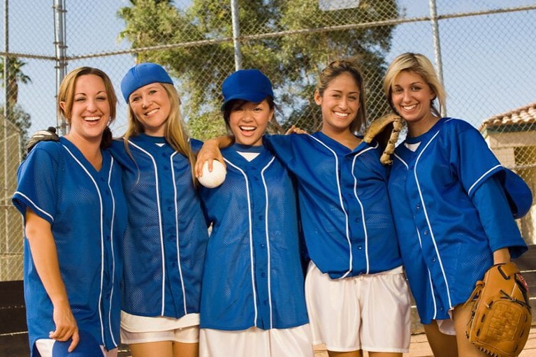 5 women in blue matching softball jerseys & white shorts pose for a picture some holding softball mits and baseball caps