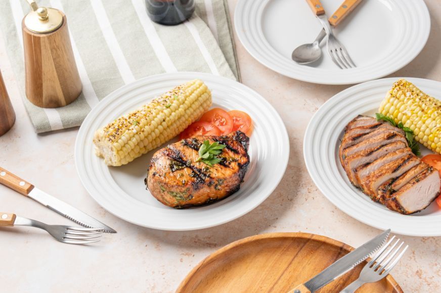 healthy summer cookout  - salmon burger, corn on the cob, sliced chicken with corn on a clean white table with white plates  silverware