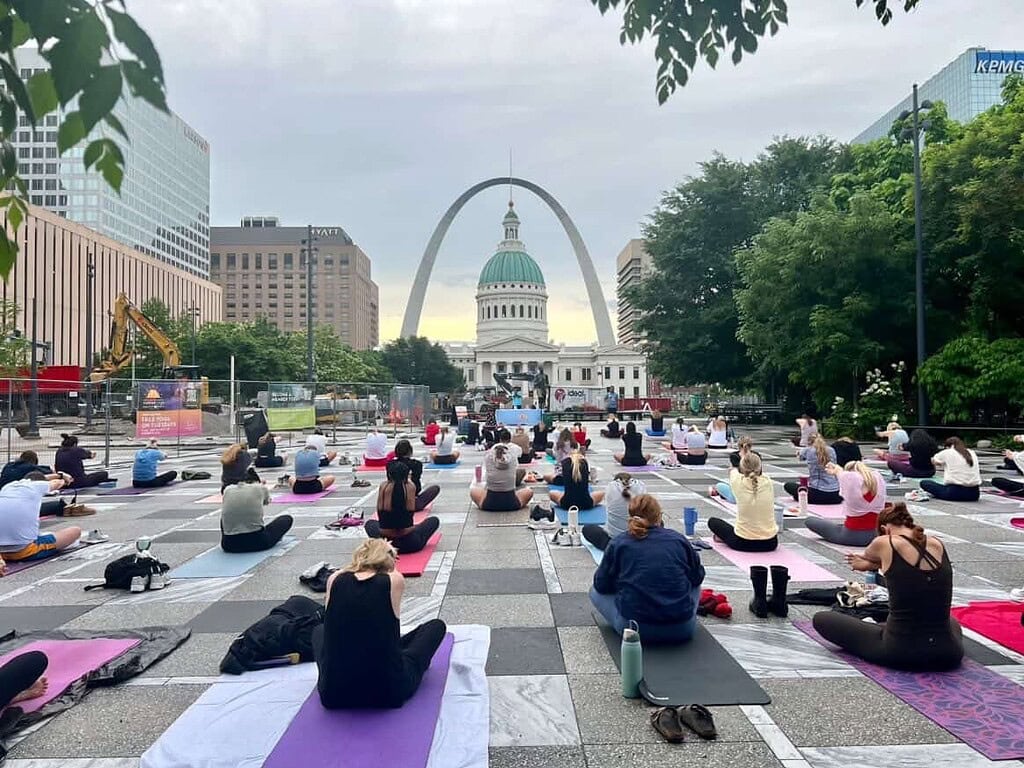 lots of yogis sitting on a their mats in front of kiener plaza with an arch view