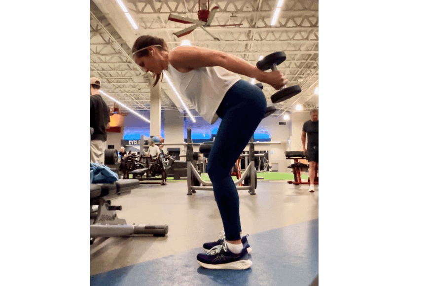 women at in the gym lifting weights behind her back, tricep exercise. she is wearing navy pants and a white shirt