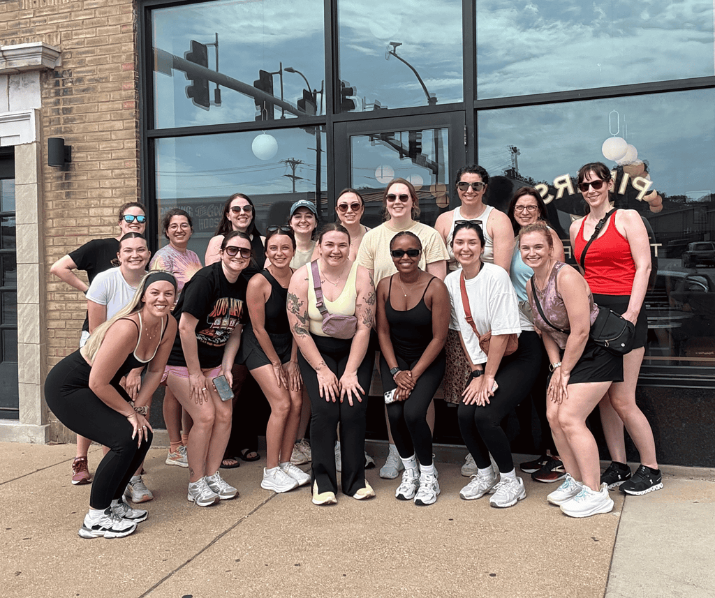 group of young women in athletic wear smiling in a group shot along a brick wall with windows