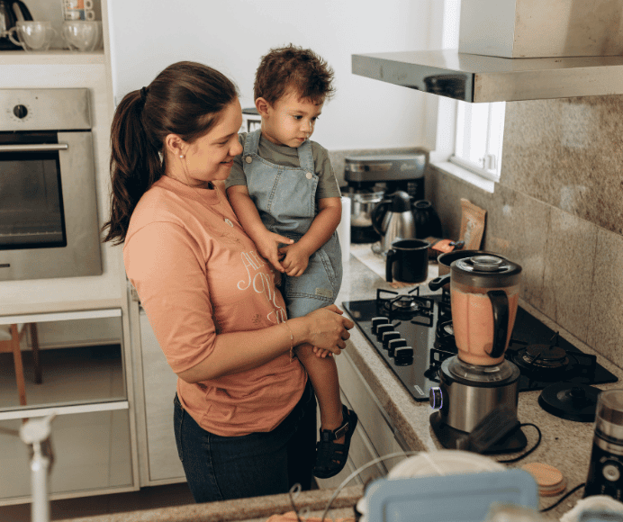 women standing in her kitchen holding her toddler son while blending a smoothie