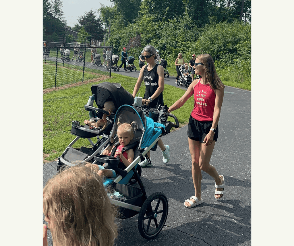 2 moms walking along a paved path on a sunny day with their strollers and and older child walking ahead