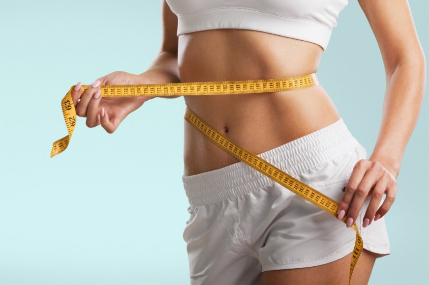 torso of a lean white woman in white shorts and white sports bra with a yellow measuring tape on her waist